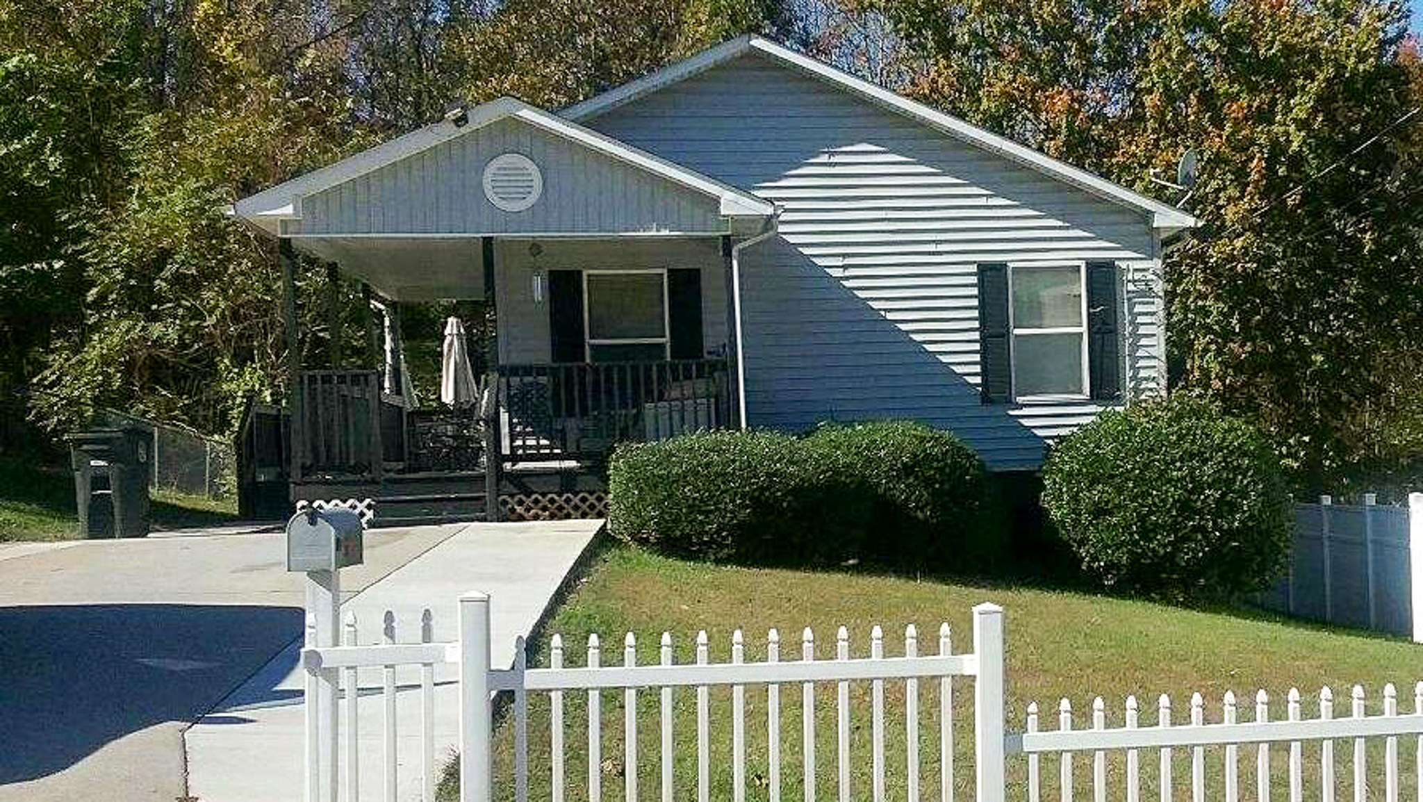 A gray house with a covered front porch, green shutters, and neatly trimmed bushes sits behind a white picket fence and a sloped driveway belonging to Sponsored Residential Provider Brenda Jackson in Lynchburg, Virginia.