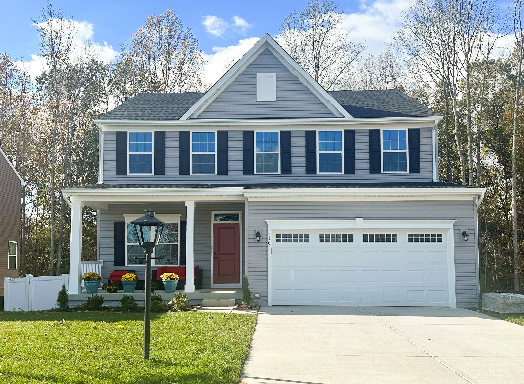 A two‑story gray house with white trim, black shutters, a red front door, and a three‑car garage, with a lamp post and fall mums in the front yard belonging to Group Home Provider Robertha Rose in Fredericksburg, Virginia.
