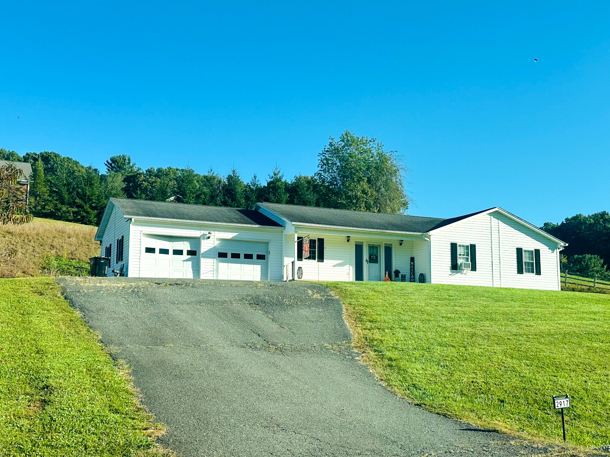 A white single‑story house with green shutters, a covered front porch, and a two‑car garage, sitting on a grassy hill belonging to Sponsored Residential Provider Jayda Utt in Galax, Virginia.
