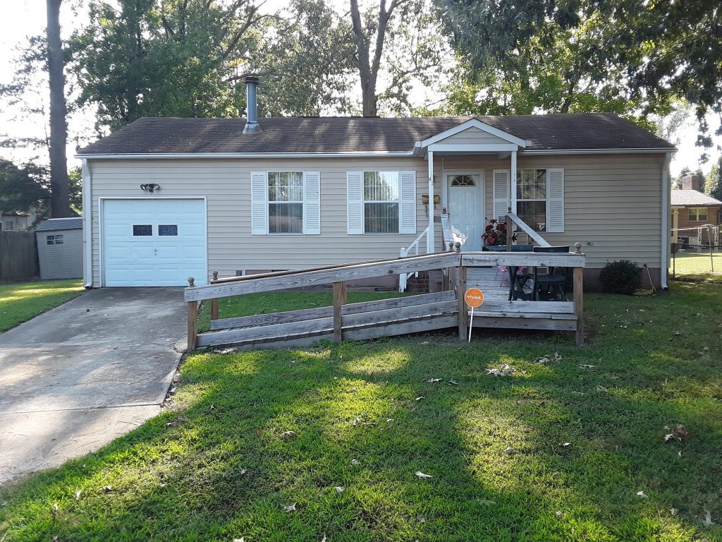 Single‑story beige house with a front ramp, small porch, attached garage, and a grassy yard shaded by trees belonging to Sponsored Residential Provider Jeanette Perry in Hampton, Virginia.