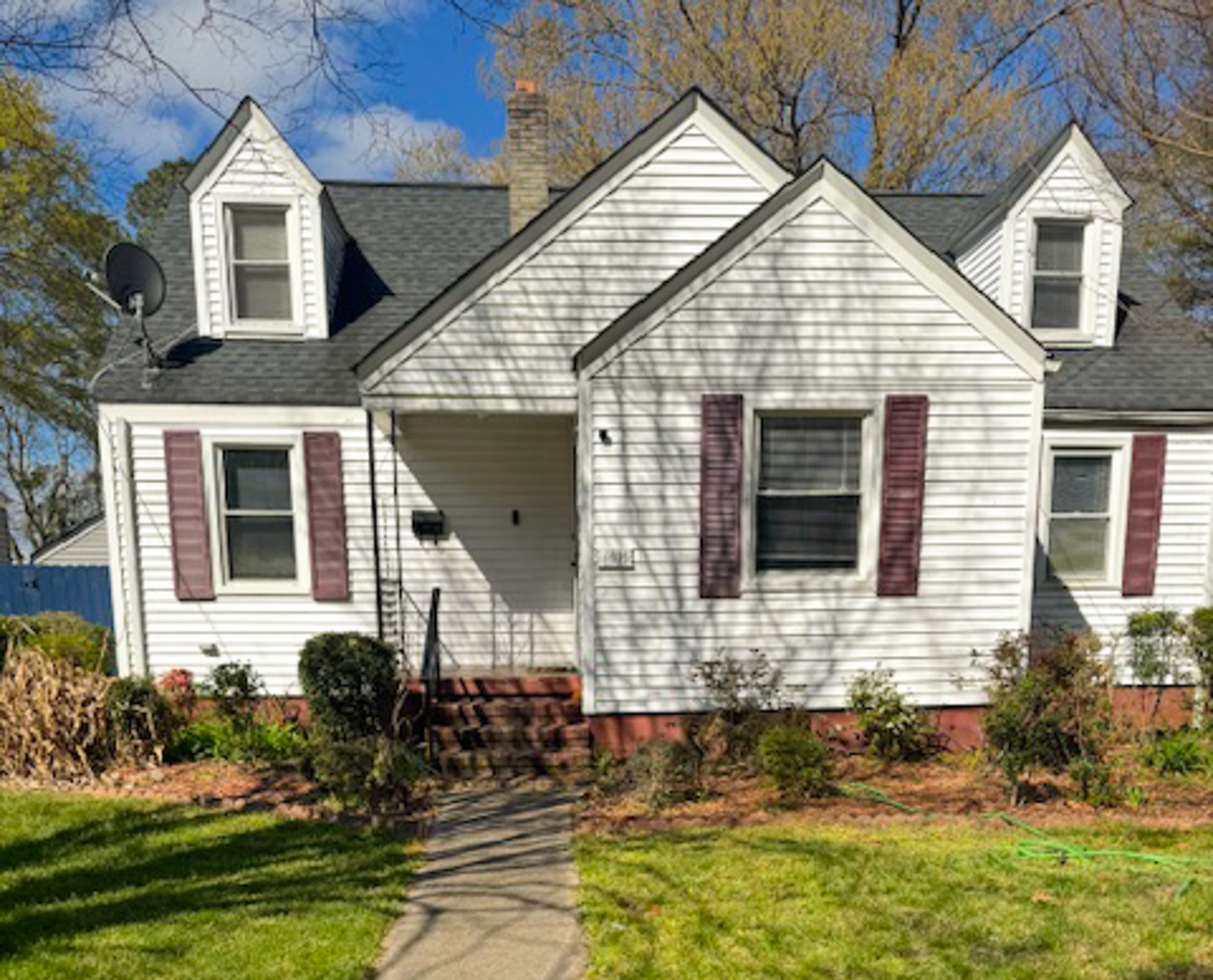 Small white house with dark shutters, a central front door, and dormer windows, surrounded by shrubs and a sunlit lawn belonging to Sponsored Residential Provider Douglas Newton in Norfolk, Virginia.