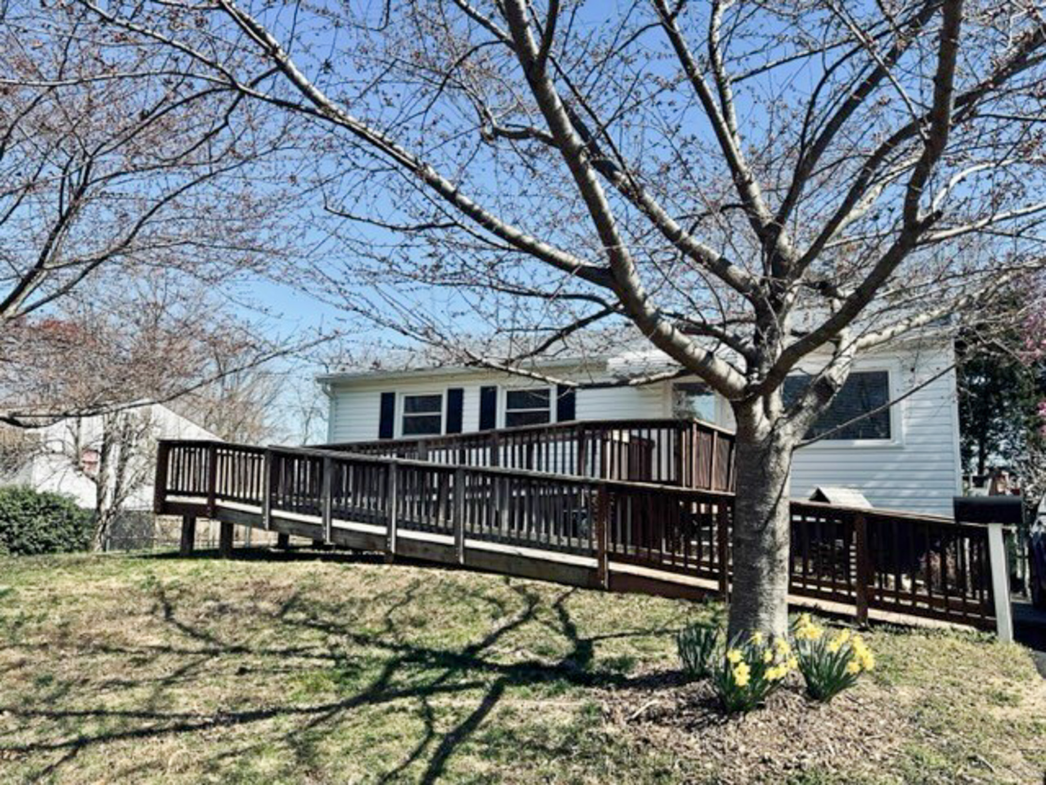 A white house with a wooden ramp in front, a large bare tree, and daffodils in the yard belonging to Sponsored Residential Provider Teresa and Bill Herran in Woodbridge, Virginia.