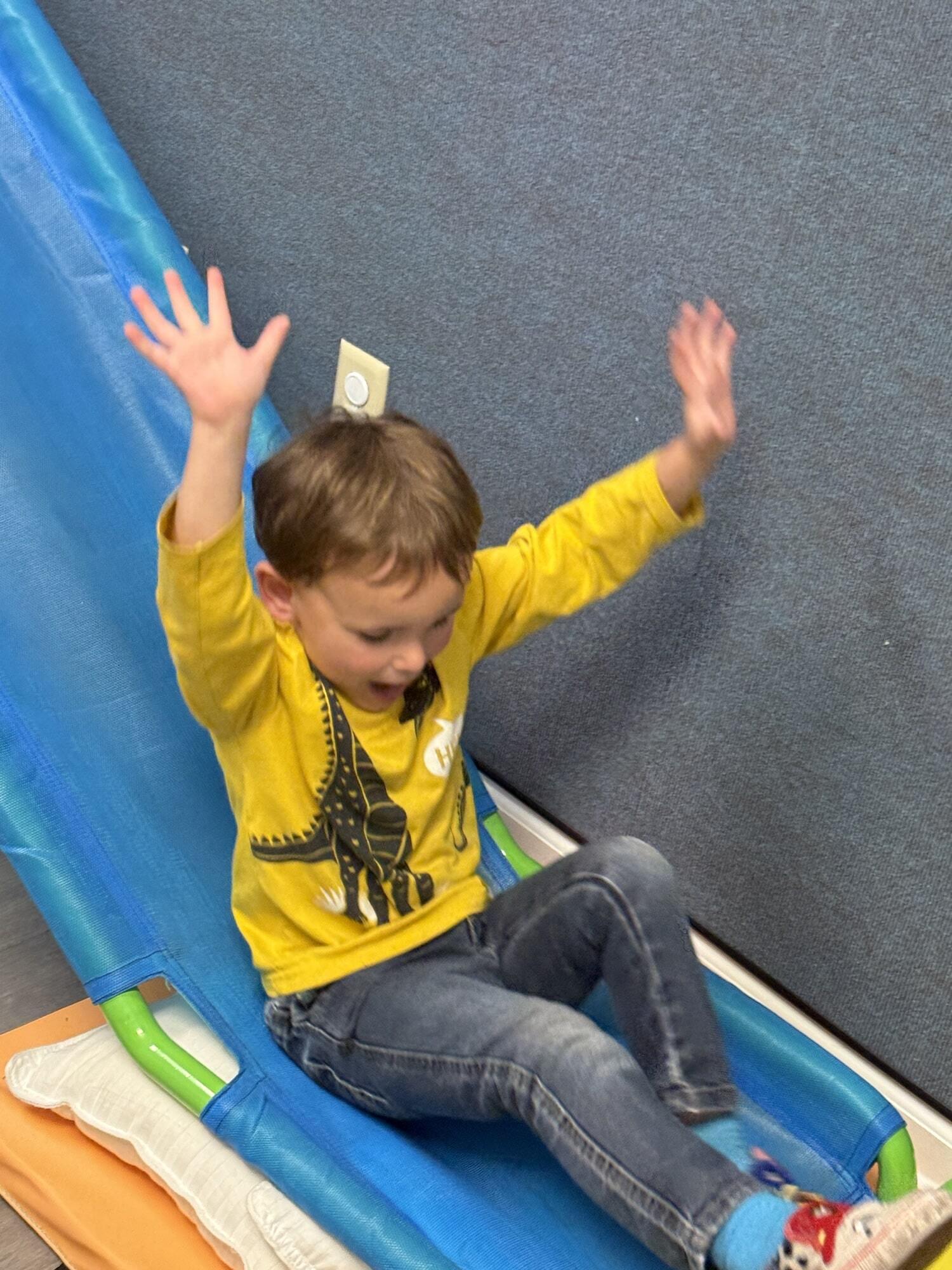 A little boy in a yellow shirt with his arms up while playing on an indoor slide during a clinic-based ABA therapy session